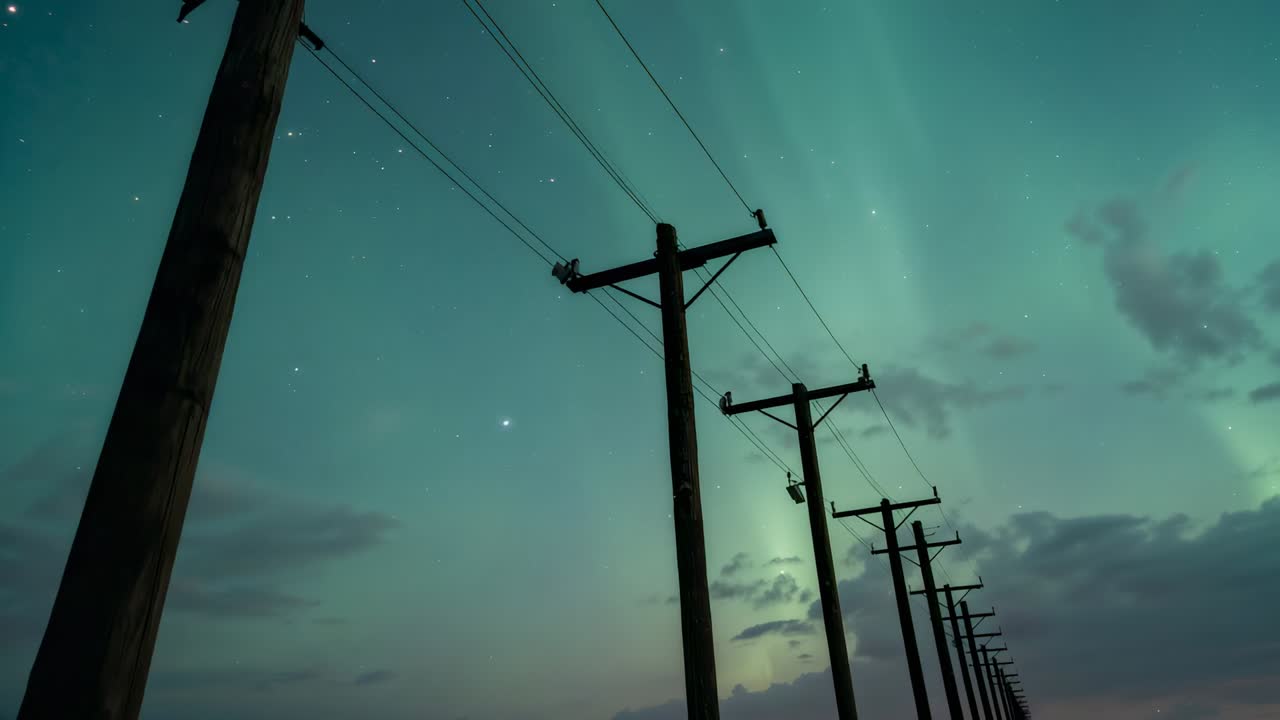 Panning camera tracing low-angle wooden utility poles and power lines at night, capturing aurora