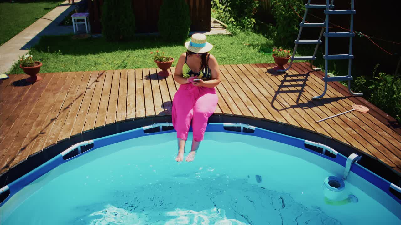 Woman in pink pants sitting on edge of swimming pool enjoying sunny day outdoors with greenery