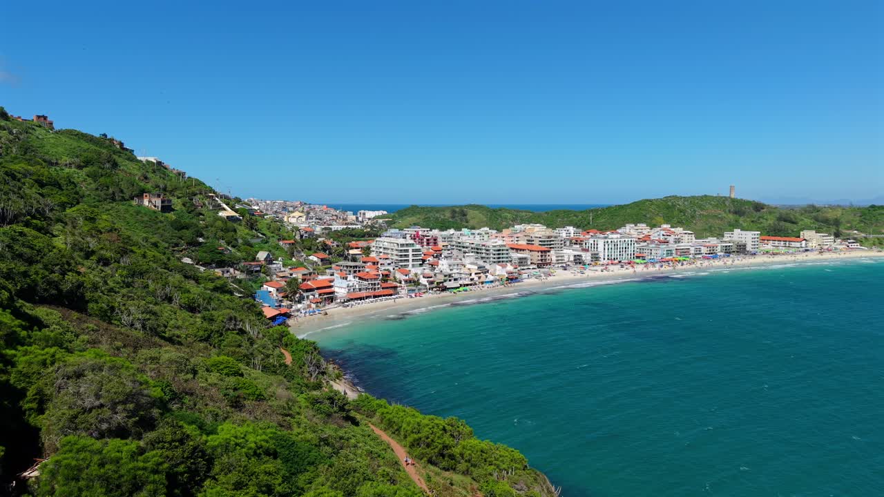 Prainha Arraial do Cabossa, Rio de Janeiron osavaltiossa, Brasiliassa, esittäen puolikuun muotoisen rannan, Atlantin rannikon ja Atlantin metsän rinteet, joista avautuu näkymä eloisalle rannikkokaupungille, lähenevä dronekuva.