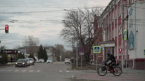 urban street view featuring a cyclist crossing at a traffic light showing a red signal and countdown, cars are lined up, and a red building is in the background