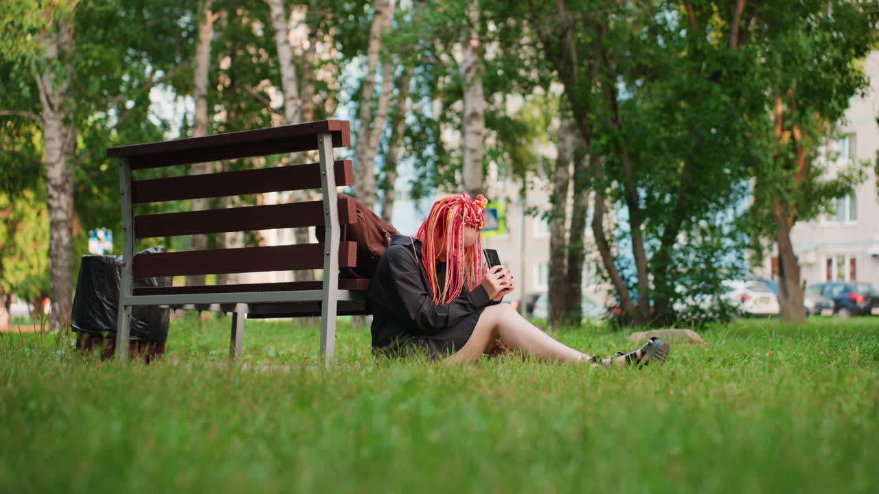 잔디 벤치에 기대어 누워 있는 화려한 드레드락을 한 백인 여성, 뒤로는 자작나무와 도시 주택이 보이고, 편안하지만 어딘가 거리가 느껴지는 자세, 조용한 그리움과 도시의 자연을 담아낸 영화 같은 넓은 뷰