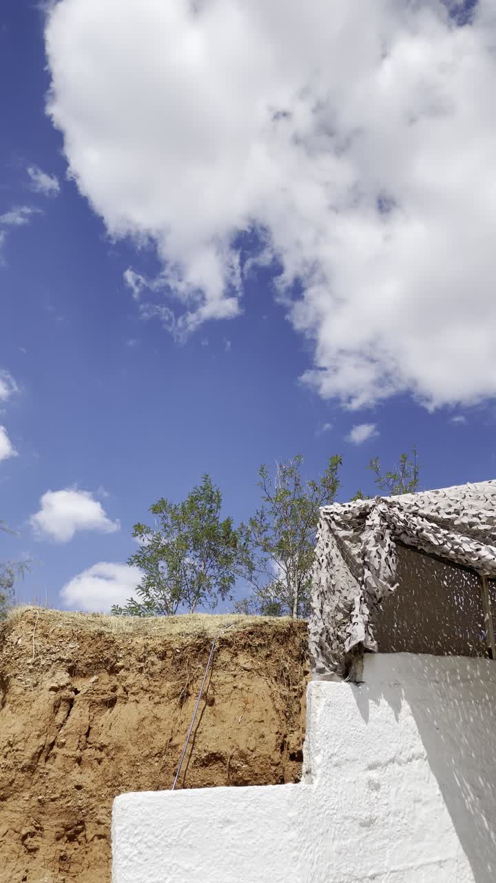 Scena all'aperto che mostra un muro di terra naturale con vegetazione sopra, una rete mimetica su una struttura bianca e un cielo azzurro brillante con nuvole sparse. Ideale per temi legati alla natura, alla sopravvivenza e alle attività all'aperto.