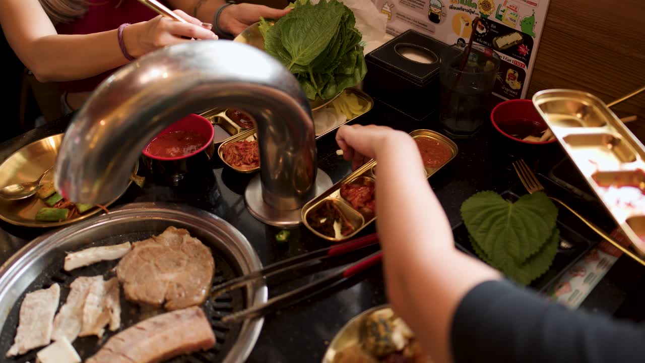 Two people grill pork belly, prepare lettuce wraps, and use chopsticks at a restaurant table
