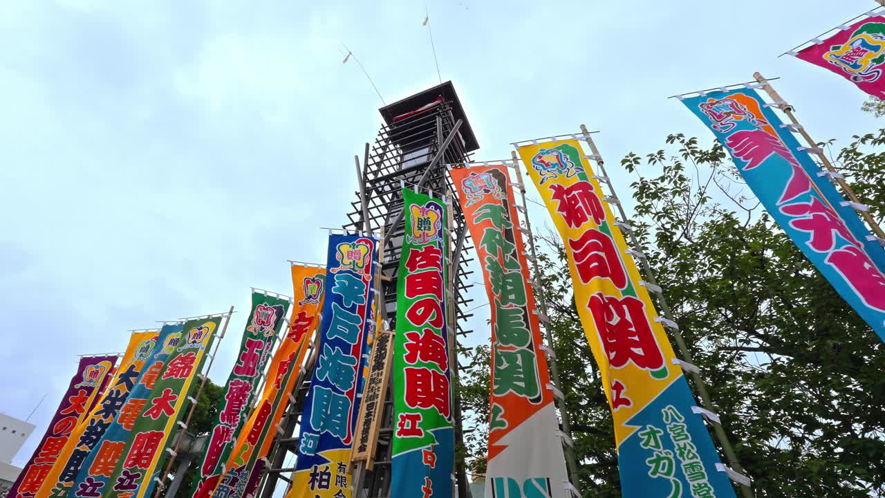 Vibrant, colorful flags (nobori) celebrating sumo wrestlers stand tall against the sky near a traditional tower, indicating a festive sumo atmosphere.