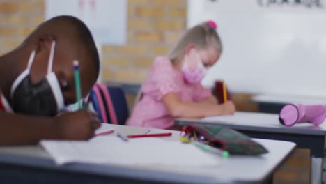 Portrait-of-african-american-schoolboy-sitting-in-classroom,-making-notes-wearing-face-mask