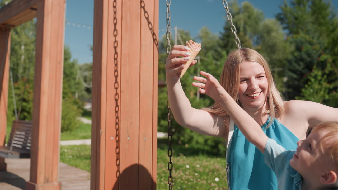 Risate di famiglia caucasica sull'altalena al parco, sorella adolescente si dondola mentre prende in giro il fratellino con un cono gelato, movimento gioioso e capelli al vento, luce solare e sfondo verde, energia spensierata del weekend
