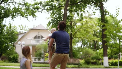 beautiful countryside with cottages on the background. green park. little blonde boy running to his father. he catch him up and throws to the air. family picnic