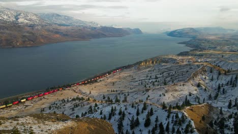 Eine-Außergewöhnliche-Aufnahme-Eines-Farbenfrohen-Zugs,-Der-Im-Winter-Am-Kamloops-see-Vorbeifährt,-Ein-Hügeliges-Wüstengelände-Mit-Fichten-Im-Vordergrund