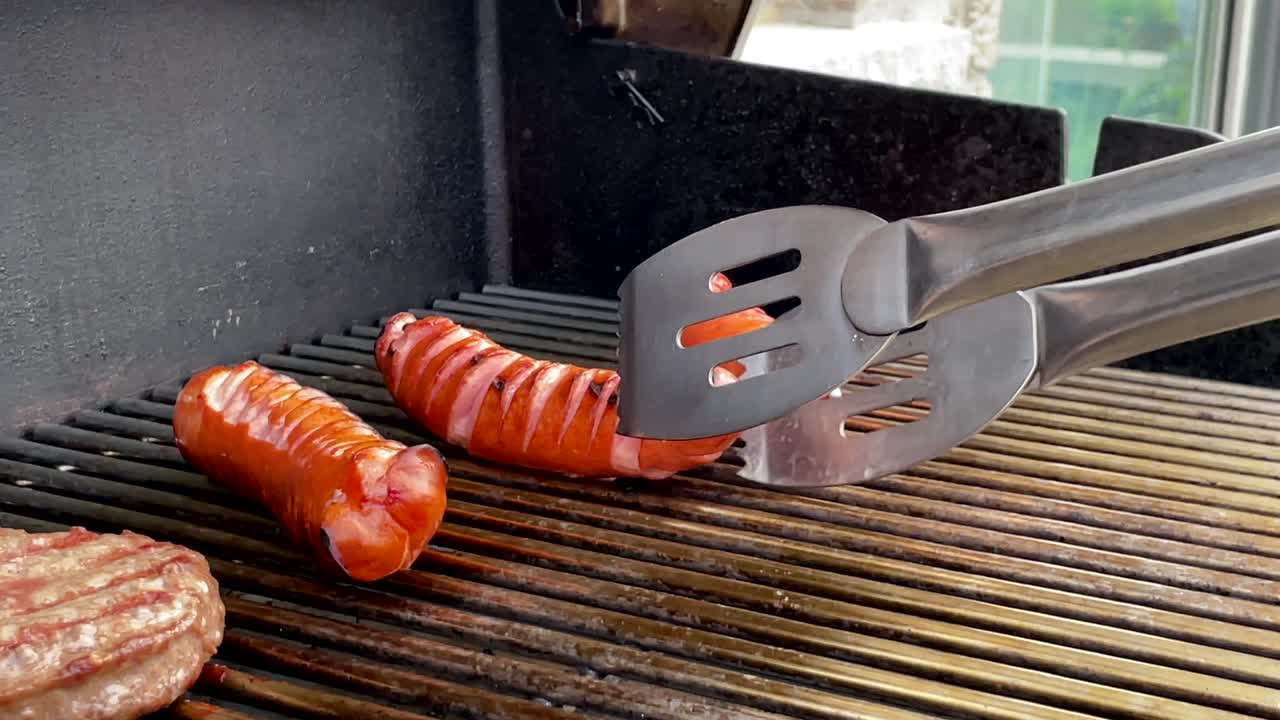 Close up of tongs flipping and adjusting hot dogs on barbecue grill, slomo