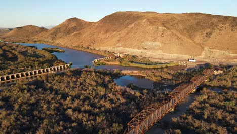 Vista-Aérea-De-La-Histórica-Represa-Y-Puente-De-Gillespie-A-Lo-Largo-De-La-Antigua-Autopista-80-Con-Vistas-Al-Río-Gila-Y-Las-Montañas-Al-Fondo