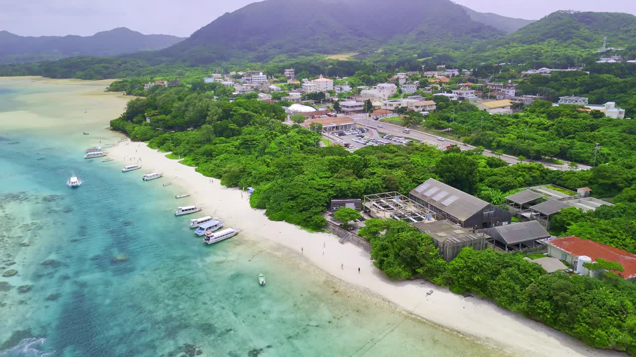 An aerial view of a boat cruising through the vibrant turquoise waters of Kabira Bay. The drone shot pans up to reveal the stunning coastline and lush, green mountains