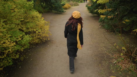 back view of a lady with a muffler around her neck and a yellow beret taking a leisurely stroll in a vibrant garden, with a blurry view of two distant figures taking another path