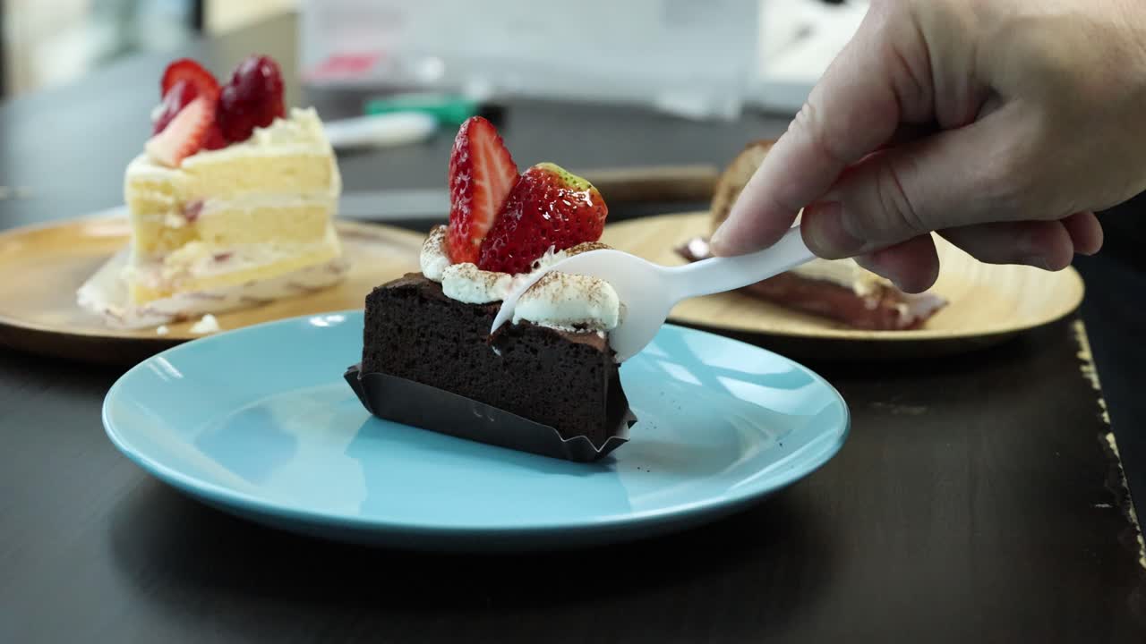 Hand uses spoon to cut and lift chocolate cake slice with whipped cream and strawberries