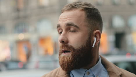 Close-up-view-of-caucasian-businessman-with-a-beard-wearing-wireless-headphones-while-talking-on-the-phone-in-the-street-in-autumn