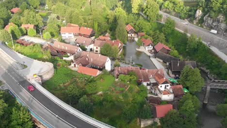 Luftaufnahme-Eines-Roten-Autos,-Das-An-Einem-Kleinen-Dorf-Vorbeifährt,-Das-Von-Wasser-Und-Wald-Umgeben-Ist