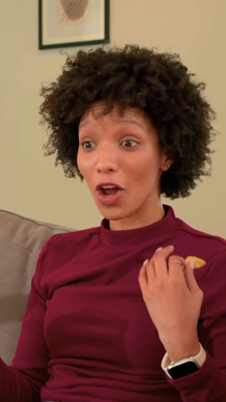 Vertical video: Talking woman in burgundy top, blue jeans with chip, prompted by companion on couch