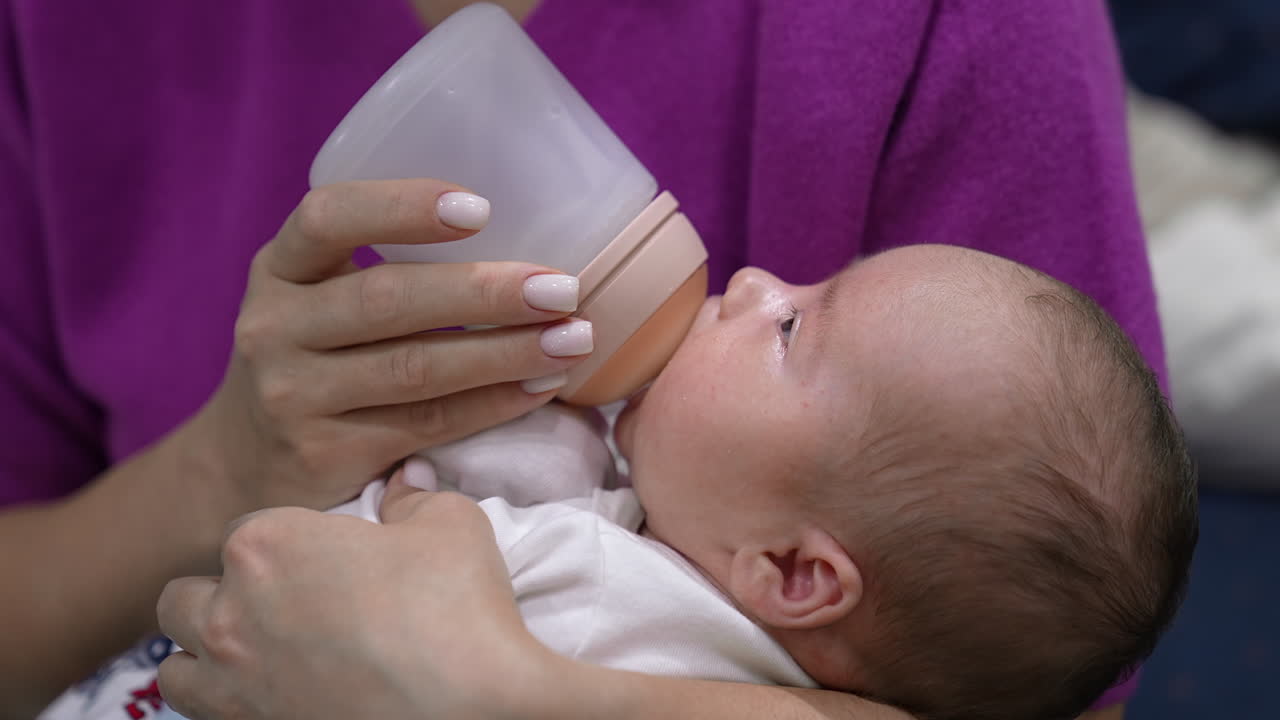Newborn kid suckling the bottle and looking up at his mommy. Cute infant in careful mother's hands being fed. Close up.