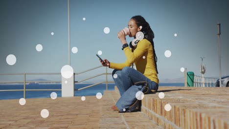 Weiße-Flecken-Auf-Einer-Afroamerikanischen-Frau,-Die-Auf-Der-Promenade-Kaffee-Trinkt-Und-Ihr-Smartphone-Benutzt