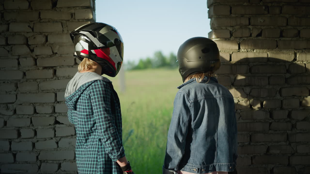 Two friends with helmets stand inside an abandoned building, gazing out towards a vast green field