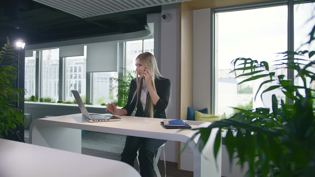 Modna bizneswoman oglądająca laptopa i rozmawiająca przez telefon. Stylowa młoda blondynka pracująca w biurze przy stole, przeglądająca laptopa i rozmawiająca przez telefon, uśmiechając się