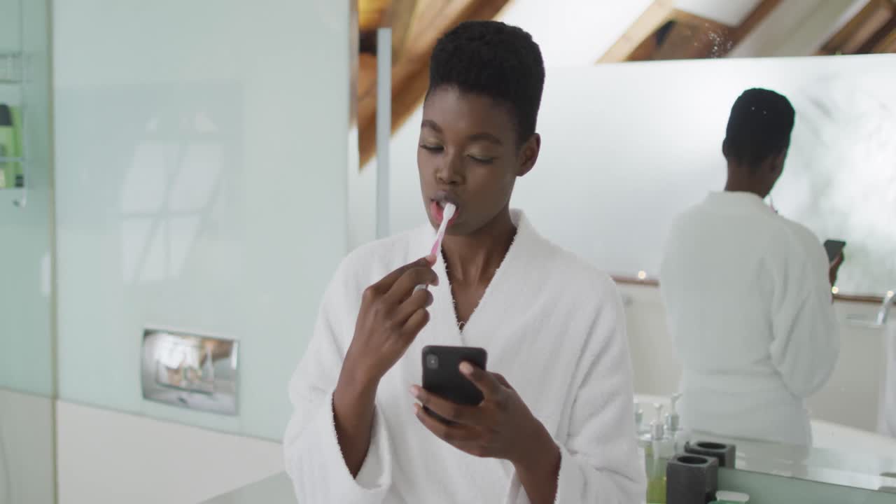 African american attractive woman brushing teeth and using smartphone in bathroom