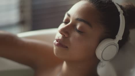 young peaceful african american woman in wireless headset relaxing in bath, enjoying water procedures at home, zoom in
