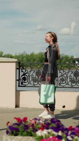 dama con ropa gris caminando al aire libre cerca de un puente, sosteniendo bolsas de compras, con un fondo borroso con una valla ornamentada, vegetación y un puente lejano