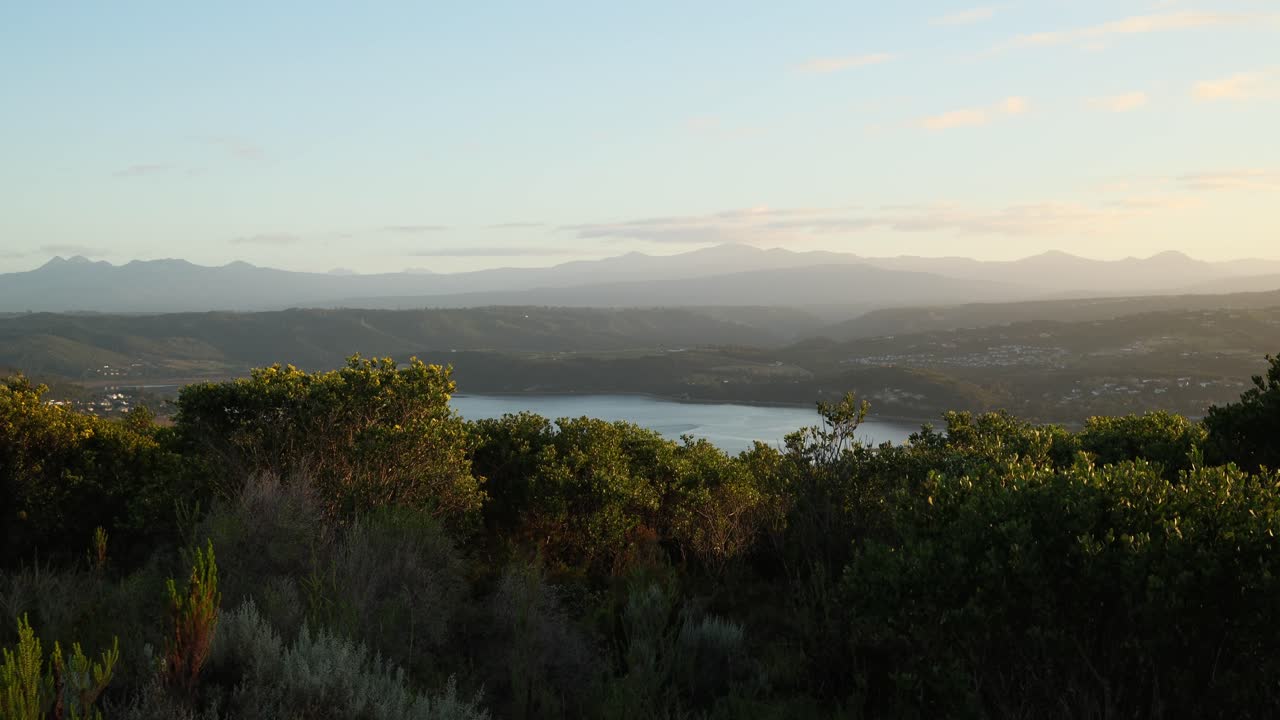 Static 4K shot of a calm lagoon framed by dense bushes in the foreground, with layered mountains in the distance. Soft morning mist drifts through the landscape under warm sunrise light