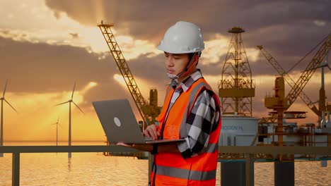 vista lateral de un ingeniero masculino asiático con casco de seguridad trabajando en una computadora portátil mientras está de pie con la producción de hidrógeno en alta mar