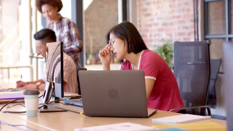 thoughtful asian woman working at computer, with diverse colleagues in casual office, slow motion