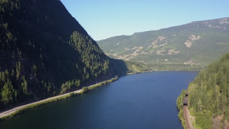 Rising-aerial:-Highway-and-railway-on-shore-of-blue-mountain-lake,-BC