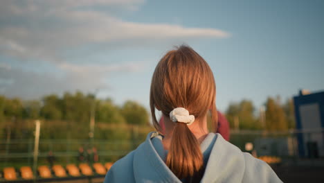 back view of lady with hair tied back playing volleyball outdoors, trees and building visible in the background, with ball in the air under clear sky and sunlight