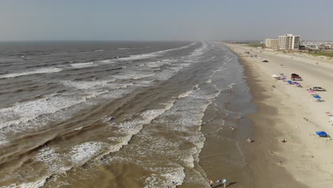 Volando-Lentamente-Por-La-Playa-Sobre-La-Línea-Divisoria-Del-Agua-Y-La-Playa-Mientras-Varias-Familias-Disfrutan-Del-Agua