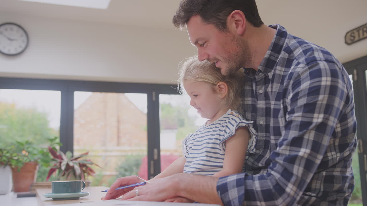 padre en casa en el mostrador de la cocina ayudando a su hija a dibujar en un libro - filmado en cámara lenta