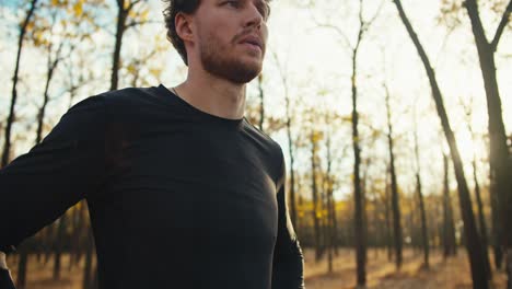 Portrait-of-a-confident-Happy-man-with-curly-hair-in-a-black-sports-uniform-and-a-beard-who-stands-in-the-middle-of-the-autumn-forest-and-looks-at-it-after-his-jog-on-a-sunny-autumn-morning