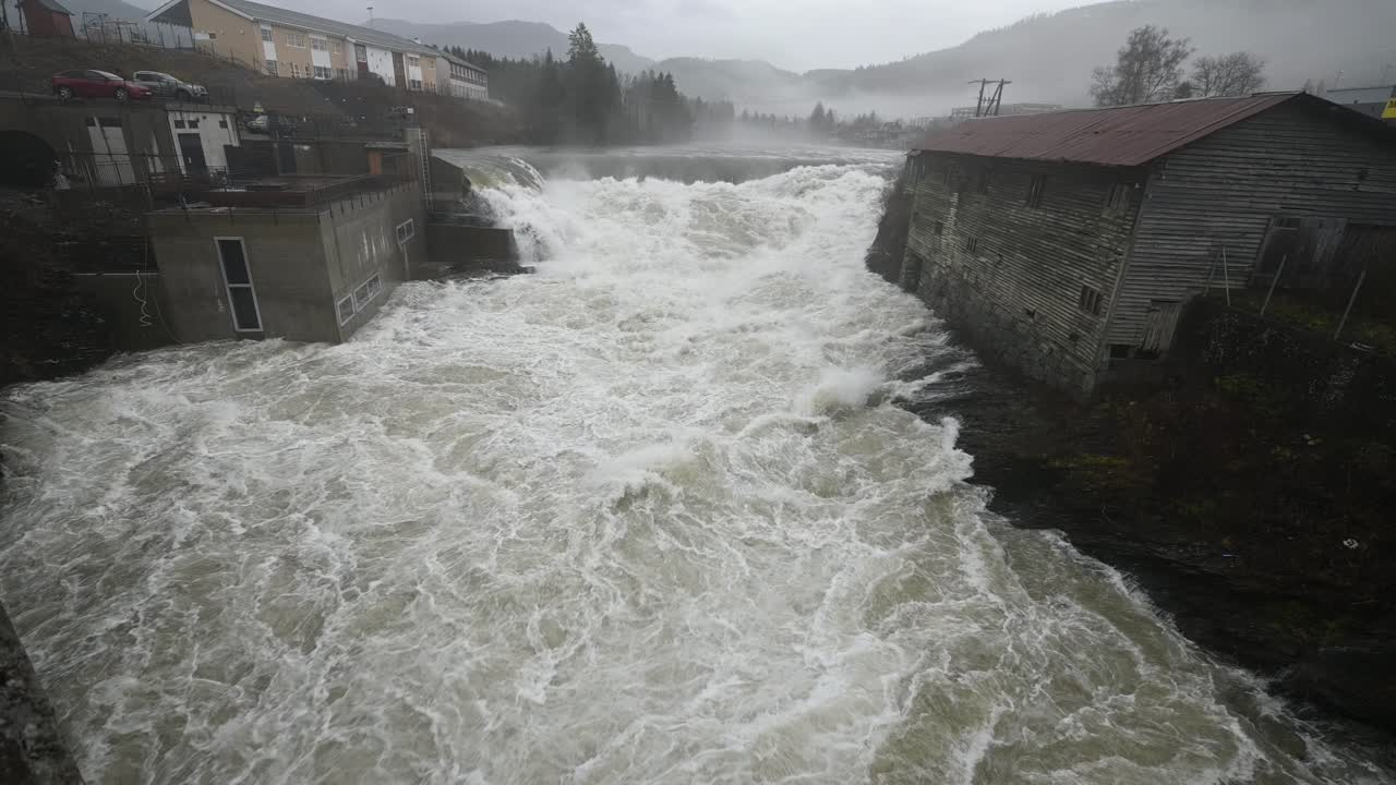 Handheld static shot of a powerful flooded river rushing under a bridge in Voss, Norway. Slow motion reveals the intense flow and energy of the water