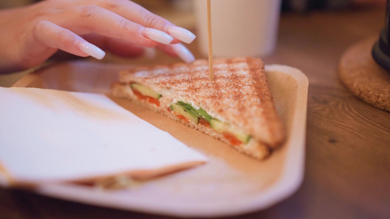 Primo piano di una mano che si protende verso un sandwich aperto su un tavolo di legno, con ingredienti visibili come cetriolo e pomodoro. La scena include un'illuminazione soffusa e texture naturali come lo sfondo con venature del legno.