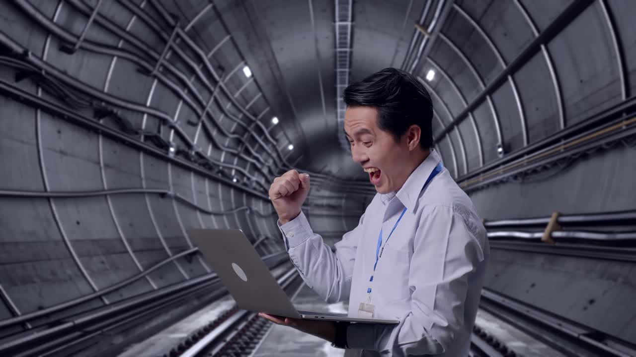 Side View Of An Asian Male Professional Worker Standing With His Laptop In Underground Subway Tunnel, He Raises His Fist Up With Screaming Goal After Typed On The Laptop