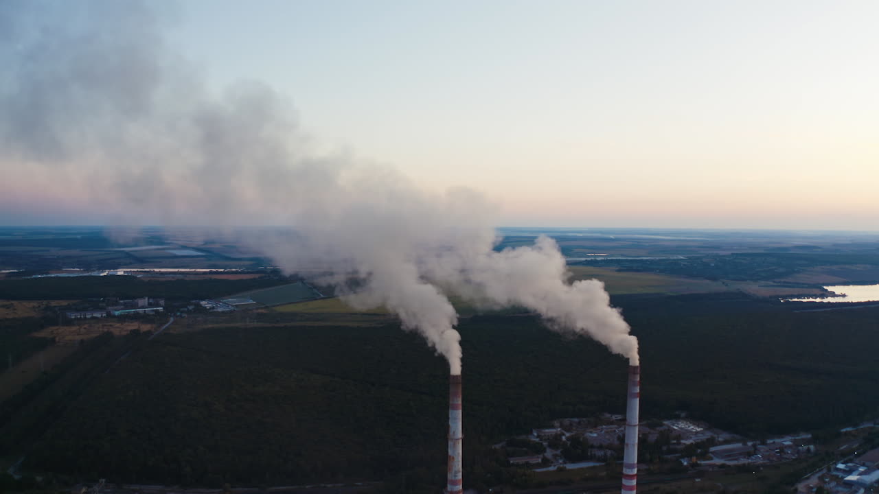 White smoke from pipes on nature background. Two chimneys produce emissions into the air among green fields and lakes.