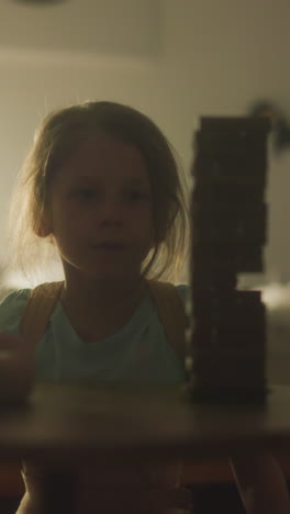 little boy gets up from table to pull block out of tower and accidentally loosens belfry. girl in background begins to worry about game. children playing board game