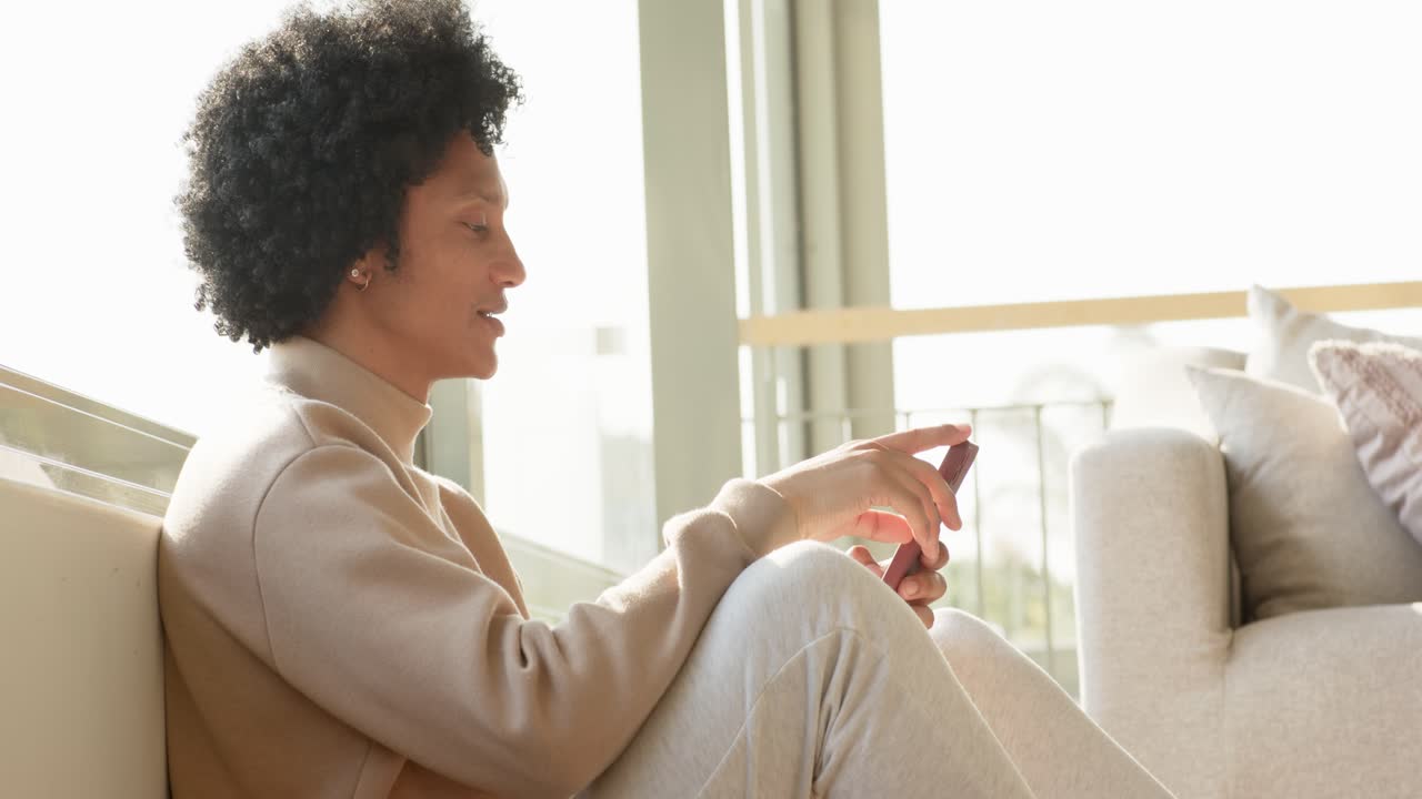Afroamerikansk mand sætter sig ved siden af sofaen på gulvet og tapper på sin smartphone, mens han kigger ud ad vinduet, kopiplads