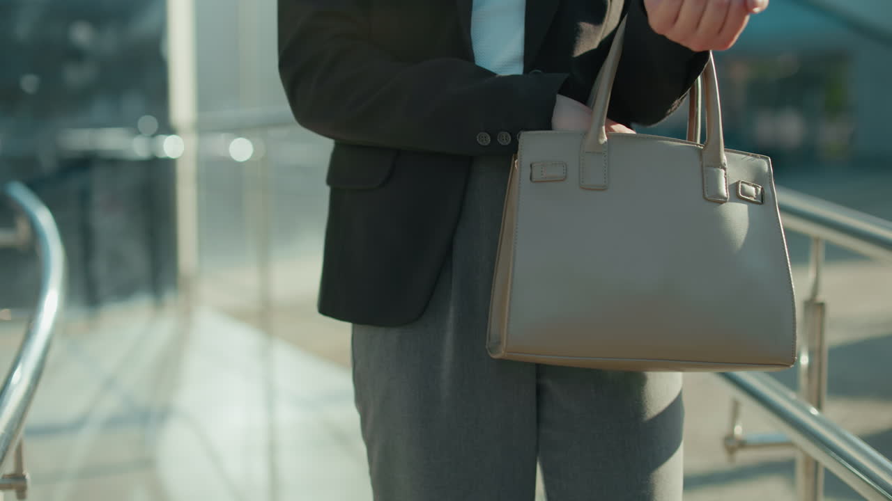 Professional lady struggles to retrieve something from handbag while getting keys, close up of elegant bag and attire, standing near metal railings in sunlit area