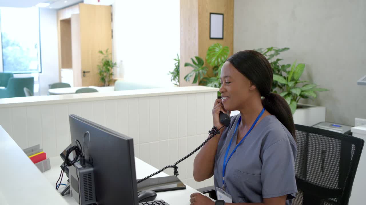Donna africana adulta in camice grigio alla reception della clinica che sente il telefono squillare, risponde al telefono e digita.