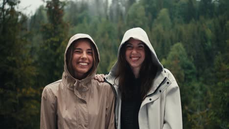 two girls in light jackets in hoods rejoice in the coming rain and look at the camera in a mountain forest