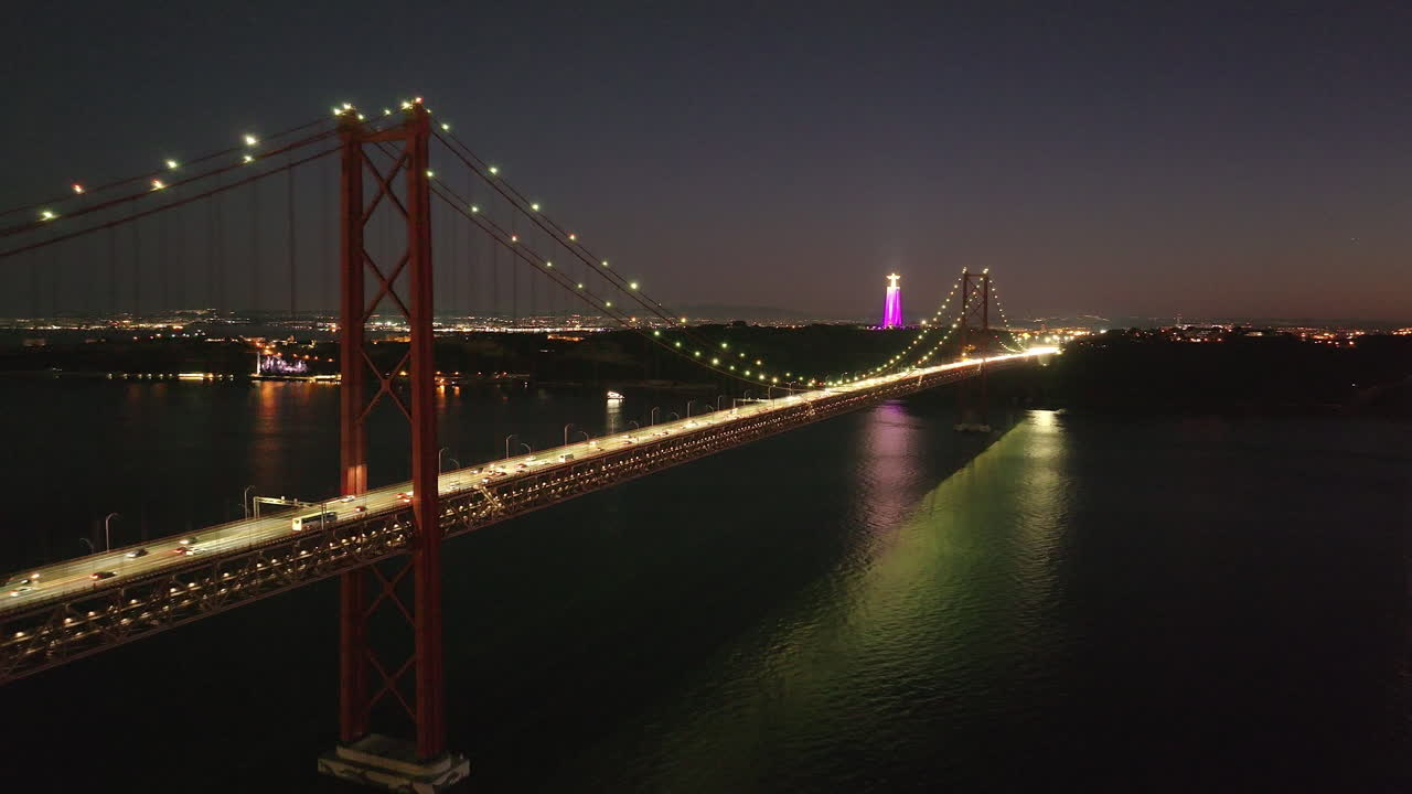 Aerial drone cinematic night establishing shot of famous red 25th April Bridge and Cristo Rei in Lisbon, Portugal, Europe. Jesus Christ statue in pink. Traffic flow, cars crossing the bridge