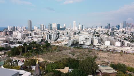 Aerial-downward-shot-revealing-a-church-steeple-in-front-of-city-buildings-view,-park-and-sky,-on-a-sunny-day