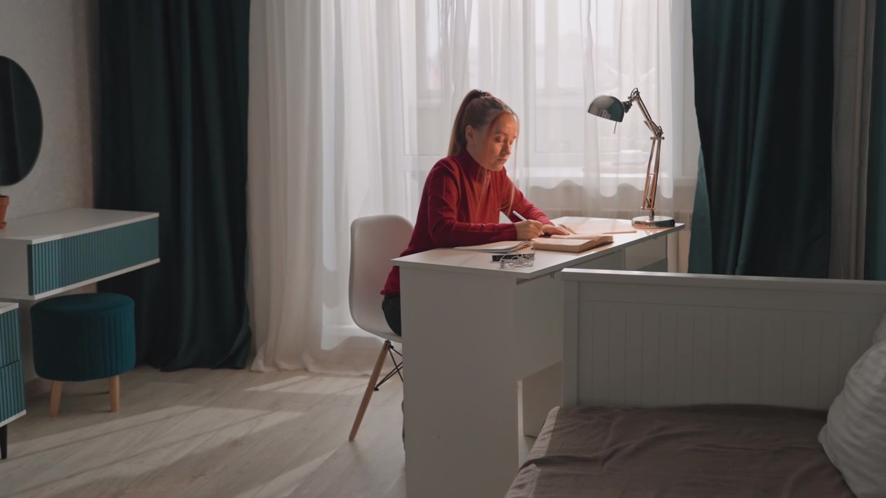 Mladá žena sedí u bílého stolu před průsvitnými záclonami. Žena si opírá pravou ruku o stů poblíž otevřené knihy. Místnost má tyrkysové stěny, sluneční světlo vrhá stíny, světlé tóny.