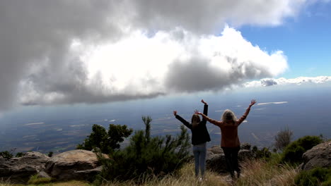 two women with hands in air enjoy view at mariepskop, from behind