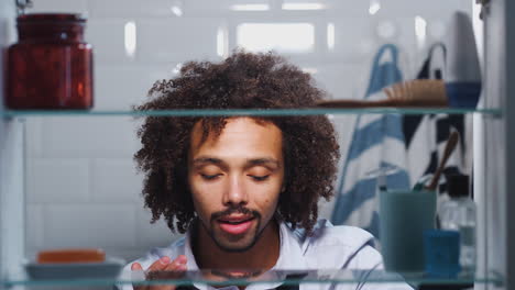 Blick-Durch-Den-Badezimmerschrank-Auf-Einen-Jungen-Mann,-Der-Sich-Auf-Die-Arbeit-Vorbereitet-Und-Seine-Haut-Mit-Feuchtigkeit-Versorgt