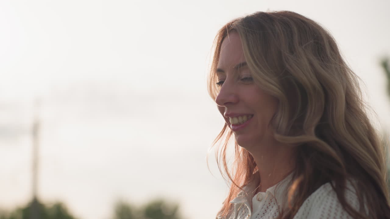 Picnic mom spreading blanket happily in sunny park, warm light on face, laughing while preparing space for family, soft background trees, casual outfit, joyful outdoor leisure moment
